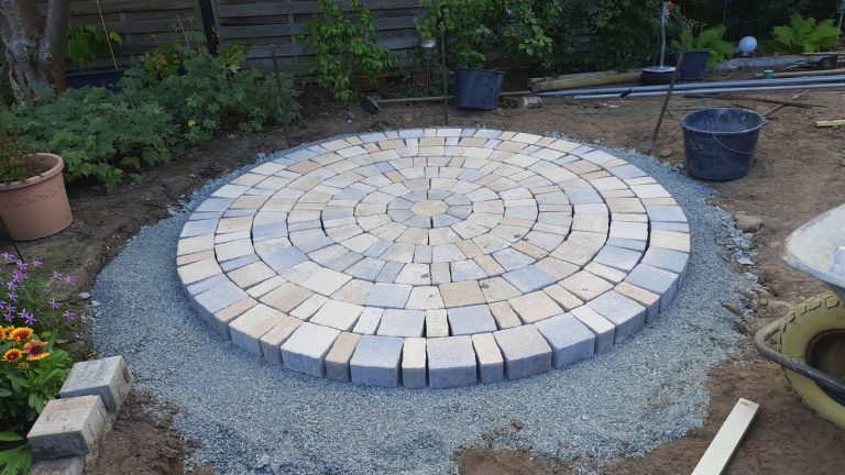 Kreisförmige Fläche aus hellgrauen und beigen Pflastersteinen, die in konzentrischen Ringen verlegt ist. Die Fläche befindet sich im Garten, umgeben von Erde, Schotter und Pflanzen.